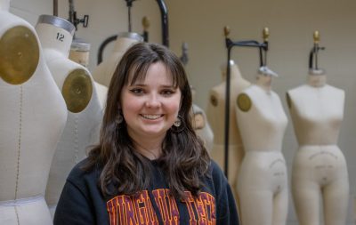 A woman with long brown hair and a gray Virginia Tech hoodie stands in front of a row of mannequins, smiling.